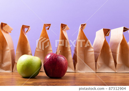 Apple and granny smith apple by row of paper lunch bags on table against purple background 87312904