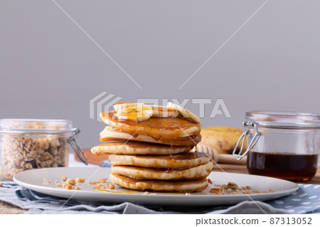Pancakes with banana slices and syrup served in plate by oats and honey jar against gray background Pancakes with banana slices and syrup served in plate by oats and honey jar against gray background 87313052