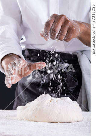 Midsection of african american male baker sprinkling flour on dough while kneading Midsection of african american male baker sprinkling flour on dough while kneading 87313259