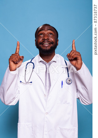 Smiling medical doctor pointing both index fingers up and wearing lab coat and stethoscope Smiling medical doctor pointing both index fingers up and wearing lab coat and stethoscope 87318737