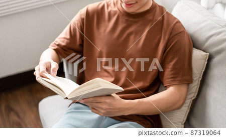 Technology Concept The male with his glasses sitting on the grey sofa, leaning his back on it, and reading a book 87319064