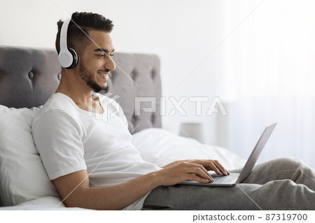 Young Arab Man Wearing Wireless Headphones Using Laptop While Relaxing In Bed Young Arab Man Wearing Wireless Headphones Using Laptop While Relaxing In Bed 87319700
