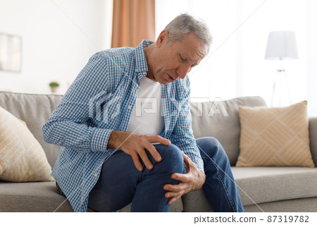 Mature man with knee pain sitting on couch at home 87319782
