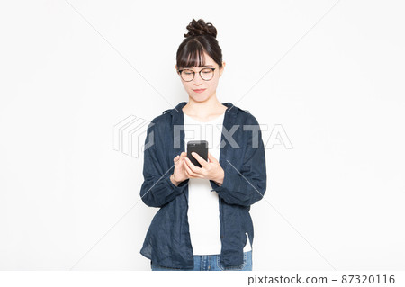 A woman wearing glasses standing in front of a white background and operating a smartphone A woman wearing glasses standing in front of a white background and operating a smartphone 87320116