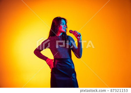 Singer. Beautiful young excited girl, student with long dark hair isolated on gradient yellow background in neon light. Concept of emotions, youth Singer. Beautiful young excited girl, student with long dark hair isolated on gradient yellow background in neon light. Concept of emotions, youth 87321153