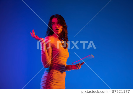 Portrait of young shocked girl, student with long dark hair using gadget isolated on dark blue background in neon light. Concept of emotions Portrait of young shocked girl, student with long dark hair using gadget isolated on dark blue background in neon light. Concept of emotions 87321177
