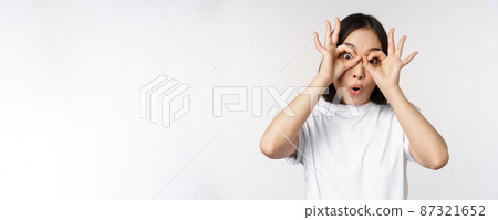 Funny young asian woman, korean girl making eyes glasses gesture, looking happy at camera, standing over white background Funny young asian woman, korean girl making eyes glasses gesture, looking happy at camera, standing over white background 87321652
