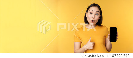 Excited asian woman showing mobile phone screen, thumbs up, like smth good, recommending smartphone application, standing over yellow background 87321745