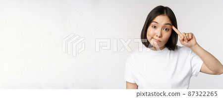 Are you crazy. Young asian woman roll finger on head, mocking someone stupid, standing in tshirt over white background 87322265