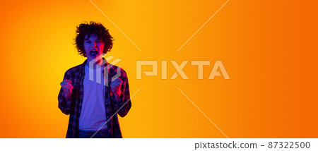 Portrait of young curly man, student in checkered shirt showing emotion of excitement isolated over orange studio background in blue neon Portrait of young curly man, student in checkered shirt showing emotion of excitement isolated over orange studio background in blue neon 87322500