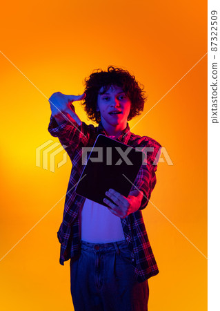 One yougn curly man in checkered shirt pointing at tablet isolated over orange background in blue neon lights One yougn curly man in checkered shirt pointing at tablet isolated over orange background in blue neon lights 87322509