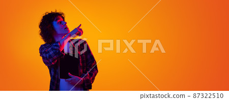 Portrait of young curly man in checkered shirt with tablet pointing right side with interest isolated over orange background in blue neon Portrait of young curly man in checkered shirt with tablet pointing right side with interest isolated over orange background in blue neon 87322510