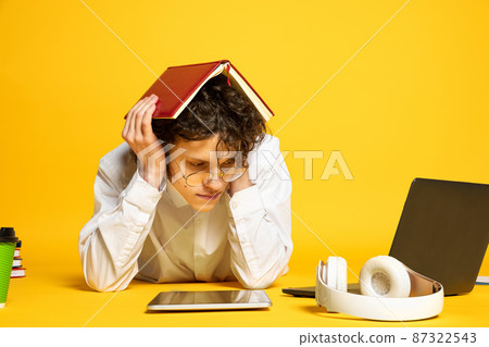 Portrait of young curly man, student in glasses lying on floor around study supplies, studying isolated over yellow studio background Portrait of young curly man, student in glasses lying on floor around study supplies, studying isolated over yellow studio background 87322543