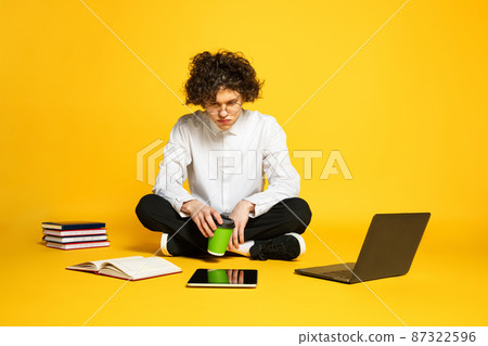 Portrait of young man, student sitting on floor, studying, preparing for exams isolated over yellow studio background 87322596