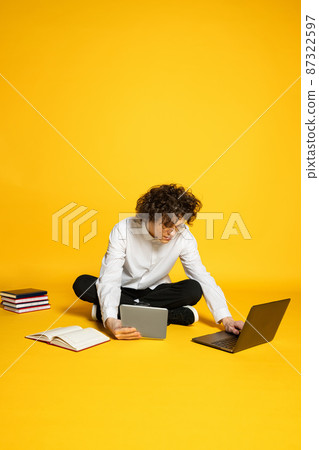 Portrait of curly young boy, student wotking on laptop, having online lessons isolated over yellow studio background Portrait of curly young boy, student wotking on laptop, having online lessons isolated over yellow studio background 87322597