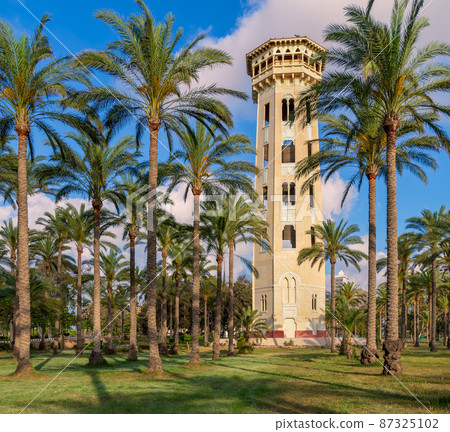 Old decorated antique elevated water tank framed by high palm trees before sunset with partly cloudy blue sky Old decorated antique elevated water tank framed by high palm trees before sunset with partly cloudy blue sky 87325102