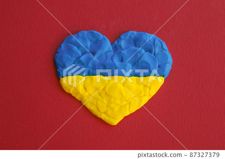Ukrainian flag. Heart shape of blue and yellow plasticine modeling clay on red maroon texture background. Plasticine finger textured background. Top view, closeup. Ukrainian flag. Heart shape of blue and yellow plasticine modeling clay on red maroon texture background. Plasticine finger textured background. Top view, closeup. 87327379