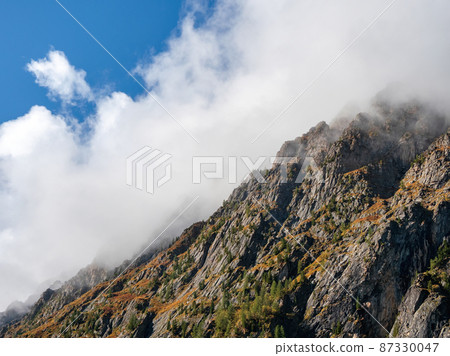 Crest of the mountain under white low clouds. Ghostly atmospheric view to big cliff in cloudy sky. Low clouds among giant rocky mountains.Natural background with high mountains, a steep mountain slope Crest of the mountain under white low clouds. Ghostly atmospheric view to big cliff in cloudy sky. Low clouds among giant rocky mountains.Natural background with high mountains, a steep mountain slope 87330047