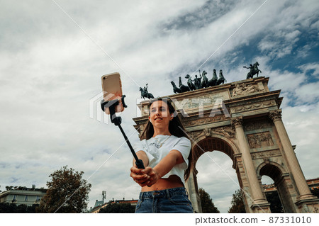 Travel blogger Milan Italy. Photographer girl with smartphone in Milan Piazza del Duomo. Paint building house in travel Europe Milano city. Hispter tourist using her phone outdoors. 87331010
