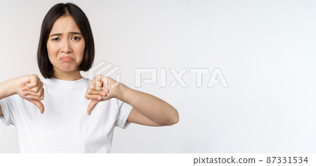 Image of asian woman showing thumbs down, dislike smth, looking disappointed, standing over white background 87331534