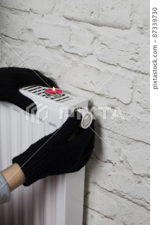 A woman's hand in winter gloves warms her hands from a heating battery. The concept of the cost of heating services, the increase in prices for utilities. Lack of heat. A woman's hand in winter gloves warms her hands from a heating battery. The concept of the cost of heating services, the increase in prices for utilities. Lack of heat. 87339730