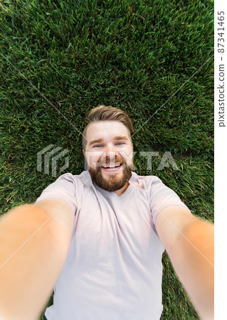 young man with beard and fashion hair style lying on grass taking selfie - holding smart phone or tablet and looking at camera. 87341465