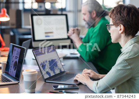 Rear view of businesswoman in eyeglasses sitting at the table and typing on laptop, she analyzing charts online with her colleagues in the background 87344243