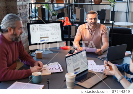 Businessman sitting at the table with computer with presentation on the monitor and listening the ideas of colleagues at meeting at office 87344249