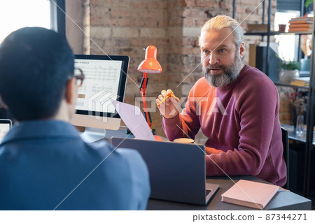 Mature bearded businessman holding document and talking about the business innovation to his colleagues at meeting Mature bearded businessman holding document and talking about the business innovation to his colleagues at meeting 87344271