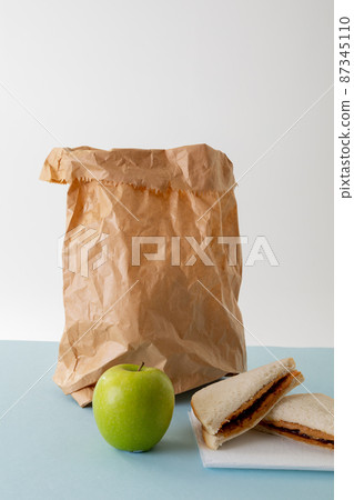 Green apple with paper bag and peanut butter and jelly sandwich on table against gray background 87345110