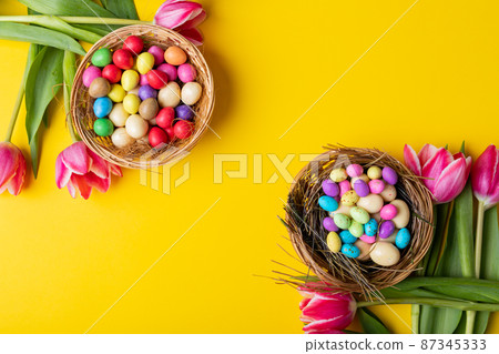 Overhead view of colorful easter eggs and candies in nests with tulips on yellow background Overhead view of colorful easter eggs and candies in nests with tulips on yellow background 87345333