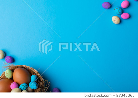 Overhead view of easter eggs with colorful candies in nest on blue background with copy space 87345363