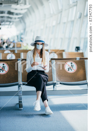 Confident business woman in a mask sitting at the airport, holding a passport and plane tickets, keeps a distance in a pandemic. Waiting at the airport. Travel concept. Woman looking at tickets. 87347793