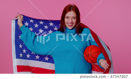 Redhead young woman basketball fan holding American USA flag doing winner gesture, dancing alone Redhead young woman basketball fan holding American USA flag doing winner gesture, dancing alone 87347807