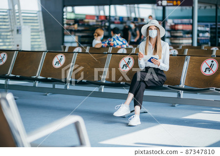 Young woman tourist wearing face mask holding passport and ticket, sitting in airport or train station on empty marked chairs under new normal and social covid distance regulations. Young woman tourist wearing face mask holding passport and ticket, sitting in airport or train station on empty marked chairs under new normal and social covid distance regulations. 87347810
