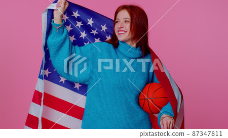 Redhead young woman basketball fan holding American USA flag doing winner gesture, dancing alone Redhead young woman basketball fan holding American USA flag doing winner gesture, dancing alone 87347811