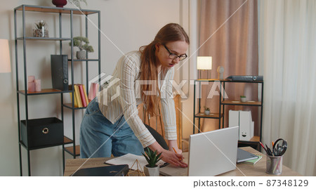 Young woman girl standing by desk in home office, working at paper documents analyzes checks data Young woman girl standing by desk in home office, working at paper documents analyzes checks data 87348129