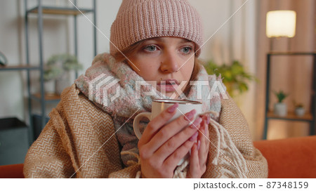 Young sick woman wear hat wrapped in plaid sit alone shivering from cold on sofa drinking hot tea Young sick woman wear hat wrapped in plaid sit alone shivering from cold on sofa drinking hot tea 87348159