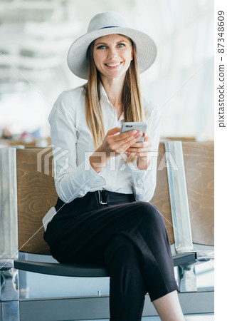 Girl uses smartphone in the airport lounge. Beautiful young girl using smartphone communication application airport arriving in new country. Woman travels around world. Smiling at the camera Girl uses smartphone in the airport lounge. Beautiful young girl using smartphone communication application airport arriving in new country. Woman travels around world. Smiling at the camera 87348609