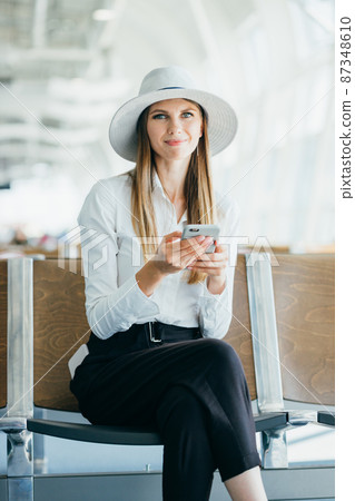 Young casual female traveler at airport, holding smart phone device, looking through the airport gate windows at planes on airport runway Young casual female traveler at airport, holding smart phone device, looking through the airport gate windows at planes on airport runway 87348610