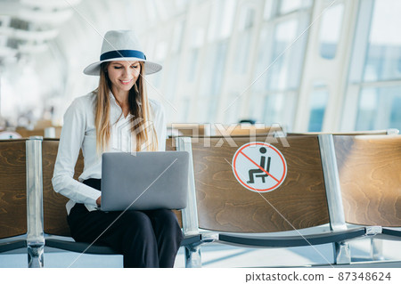 Departure lounge with a woman typing on a laptop. Girl tourist freelancer works and waits for flight in waiting room. Concept travel, remote work. Silhouette against the background of a large window. 87348624