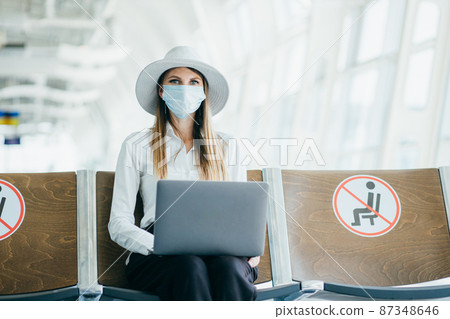 Woman in medical mask sitting at the airport terminal with a laptop. Young girl is waiting for departure at the gate and working online. Woman in medical mask sitting at the airport terminal with a laptop. Young girl is waiting for departure at the gate and working online. 87348646