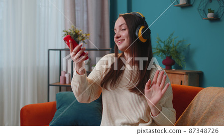Overjoyed young woman in wireless headphones dancing, singing on cozy couch in living room at home Overjoyed young woman in wireless headphones dancing, singing on cozy couch in living room at home 87348726