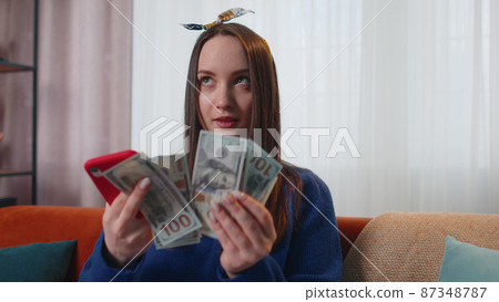 Smiling happy young woman holding cash money and mobile phone calculate domestic bills at home Smiling happy young woman holding cash money and mobile phone calculate domestic bills at home 87348787