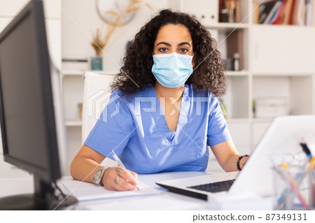 Female therapist in protective mask working at table with laptop in office 87349131