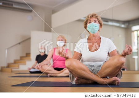 Group of aged women in protective masks, perform an exercise while sitting in the half-lotus position Group of aged women in protective masks, perform an exercise while sitting in the half-lotus position 87349418