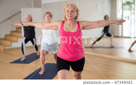 Three mature women perform the exercise by taking the warrior pose II Three mature women perform the exercise by taking the warrior pose II 87349581
