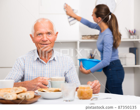 Elderly man at tea table, young woman cleaning furniture at home Elderly man at tea table, young woman cleaning furniture at home 87350135