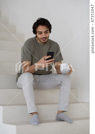 Young modern manager in casual clothes sitting on white staircase and texting 87351047