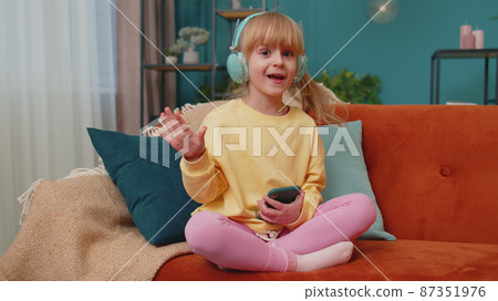 Child girl kid with smartphone in headphones dancing singing listening music at home alone on sofa 87351976